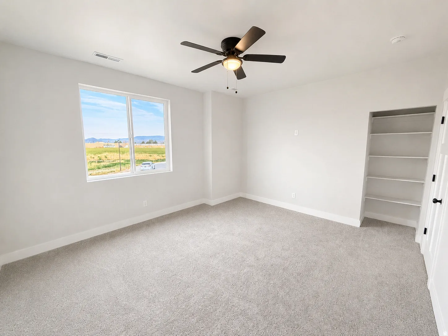 Light grey carpet flooring, white walls, built-in shelving, black ceiling fan with integrated lighting, and a large window