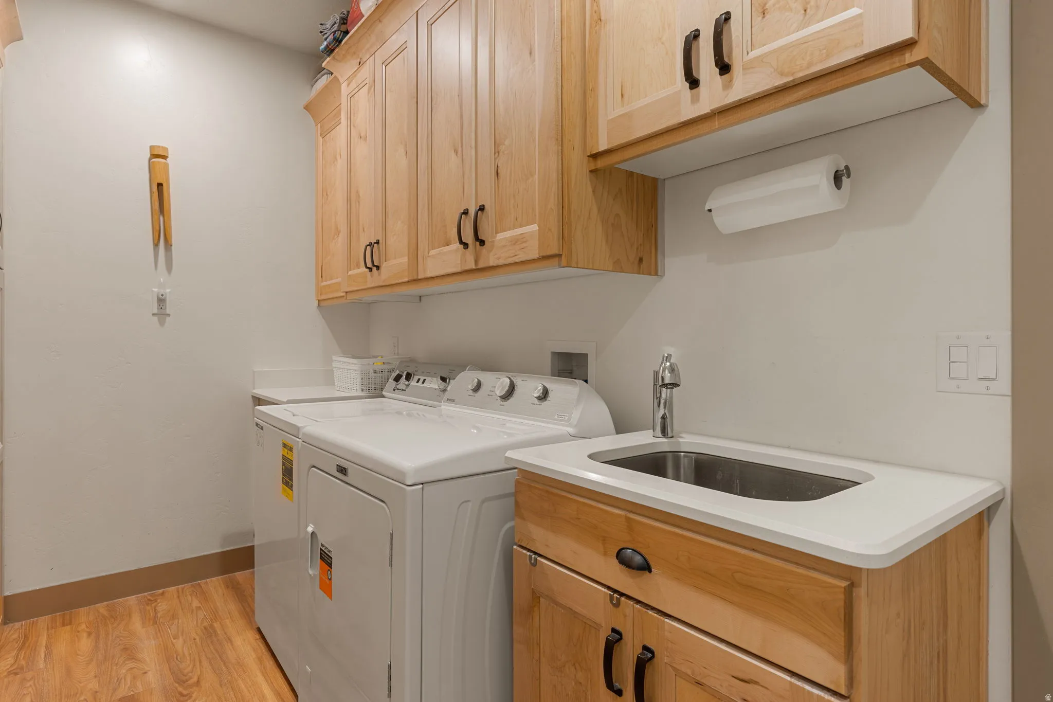 Light wood-finish cabinetry with dark hardware, white countertop, stainless steel utility sink with a chrome faucet, and wood-finish flooring