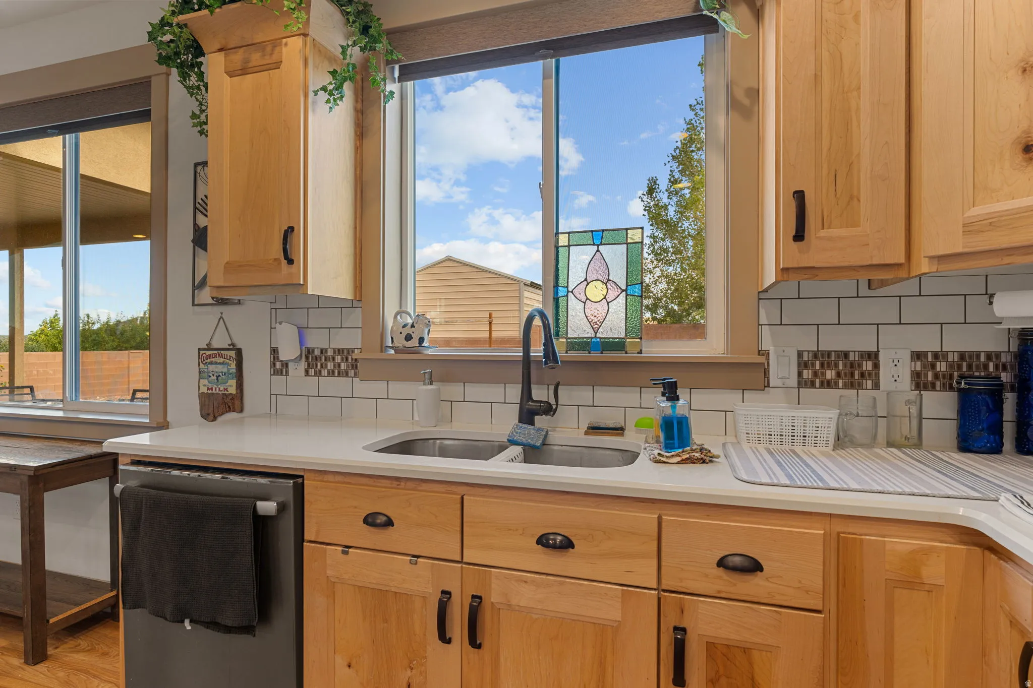 Hardwood cabinetry with dark hardware, white countertop, stainless steel sink with a dark faucet, white subway tile backsplash with accent tile, and a built-in dishwasher