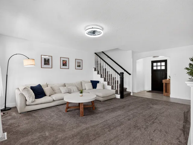 Spacious living area featuring neutral carpet flooring, a contemporary ceiling fan, white walls, and a staircase with dark wood accents