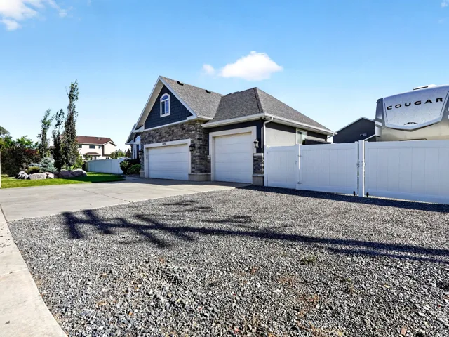Exterior featuring stone and siding finishes, a two-car garage, a gravel parking area, and a white privacy fence with double gates