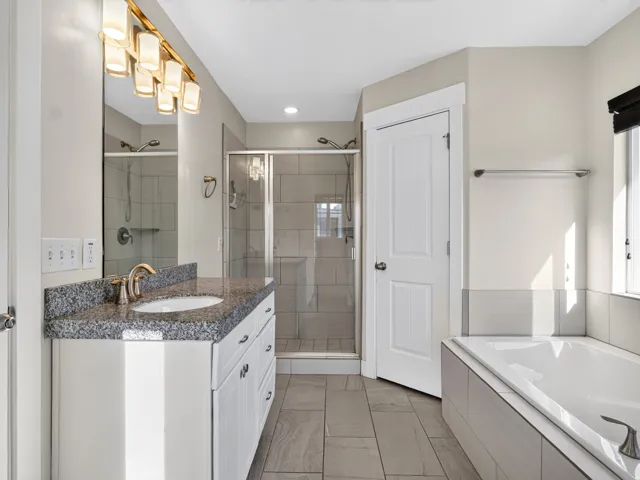 Spacious bathroom featuring a built-in soaking tub, glass-enclosed shower, single vanity with granite countertop, and tiled flooring