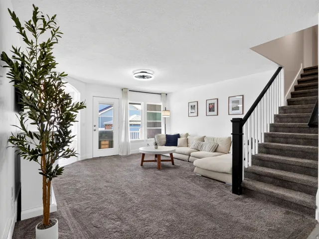Spacious living area featuring neutral carpeting, white walls, a staircase with a black handrail and white spindles, and a white exterior door with glass panels