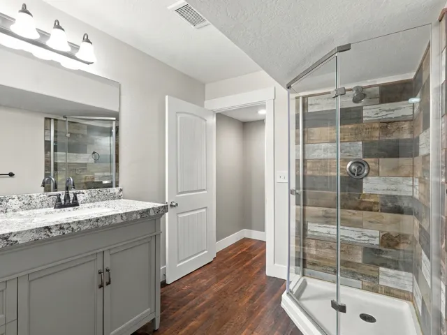 Bathroom featuring a glass-enclosed shower with decorative wall tile, a single vanity with stone countertop, and wood-finish flooring