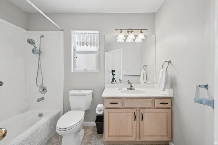 White tub and shower combination with a handheld showerhead, light wood-finish vanity with a white countertop and integrated sink, and a multi-bulb vanity light fixture