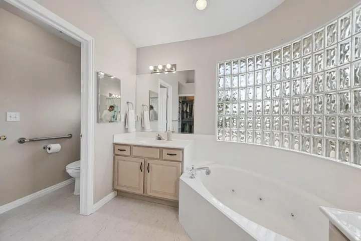Soaking tub with a curved glass block wall, vanity with a light countertop, and recessed lighting