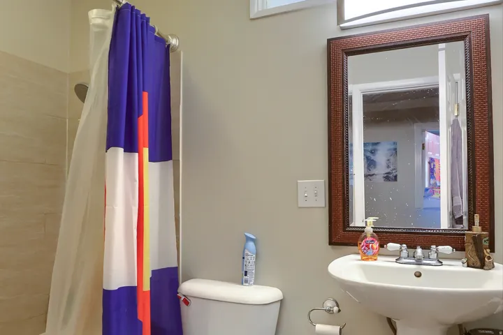 Pedestal sink with chrome faucet, framed mirror, wall-mounted light fixture, and tiled shower surround