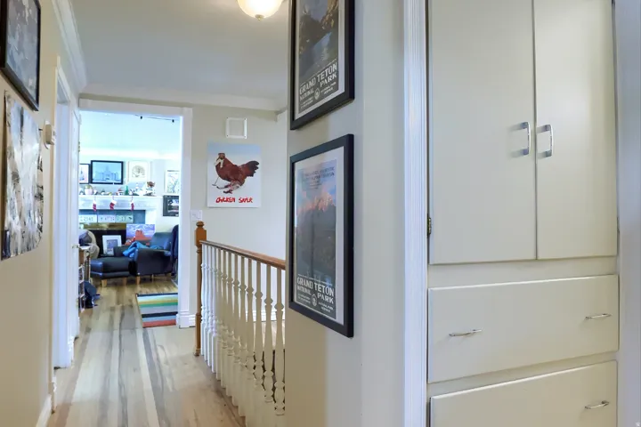 Hallway featuring wood-finish flooring, white painted walls, built-in storage cabinetry with polished hardware, and a staircase with a white railing and wood handrail