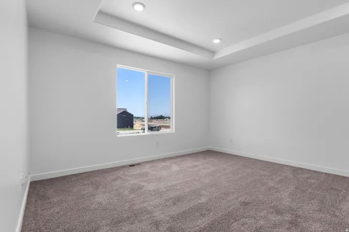Neutral tone carpeting, tray ceiling with recessed lighting, and a large window