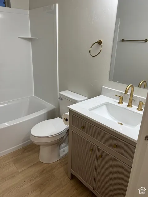 Wood-finish flooring, white bathtub with a built-in shelf, white toilet, vanity with a white countertop and integrated sink, and brass fixtures