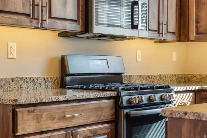 Granite countertops with an integrated backsplash, dark wood cabinetry, stainless steel microwave, black gas range, and under-cabinet lighting