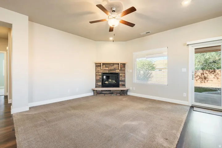 Living area featuring a stone-clad corner fireplace with hearth, a ceiling fan with integrated lighting, a sliding glass door providing exterior access, and wood-finish flooring extending into an adjacent space