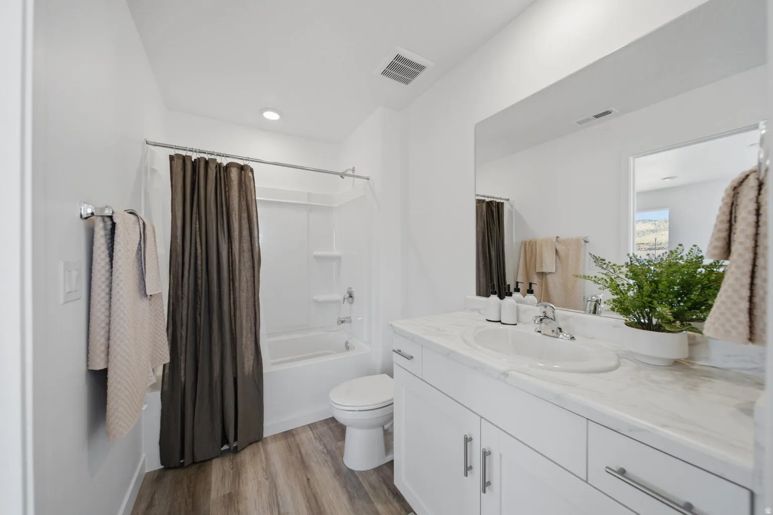 White vanity with integrated sink, wood-finish flooring, full-width mirror, and a white tub with shower