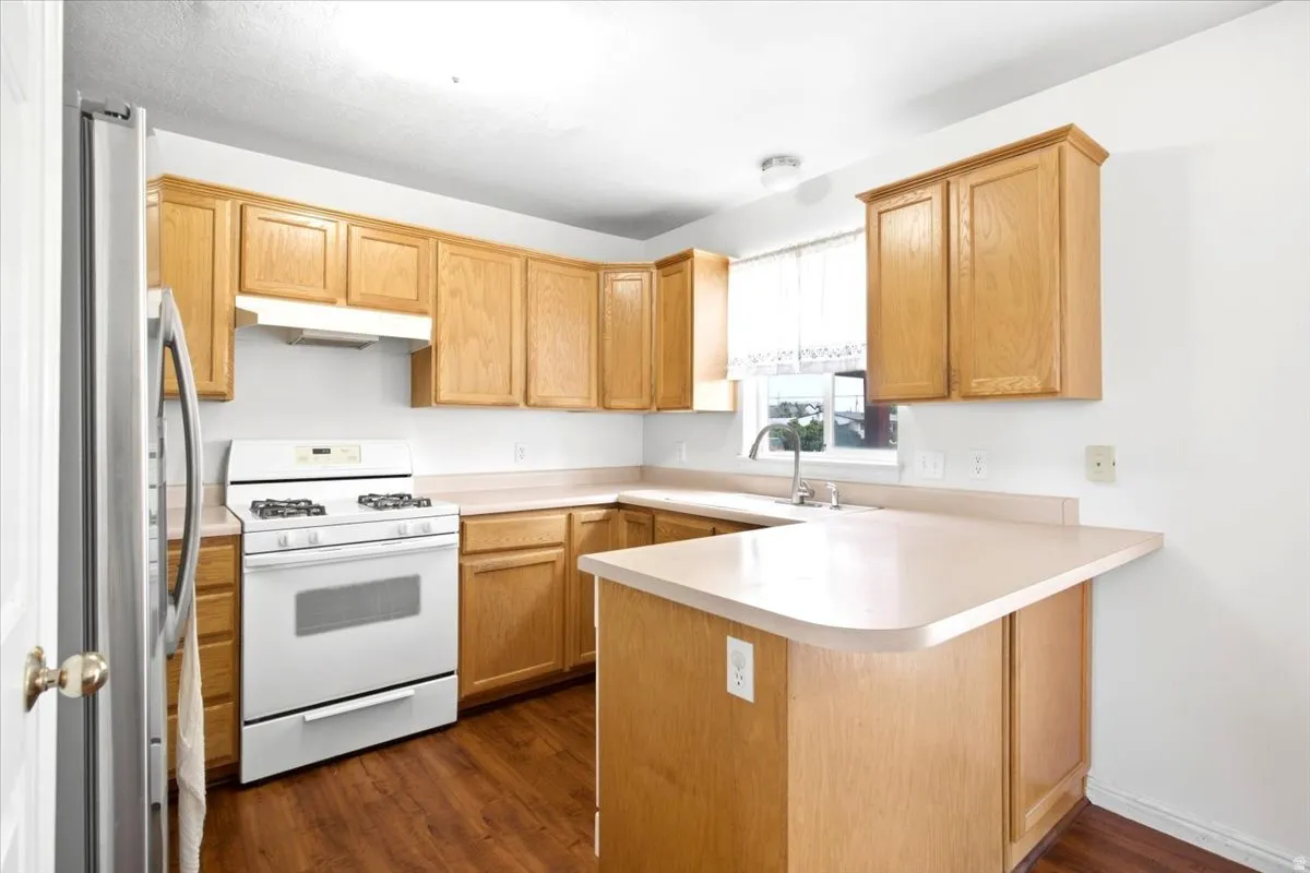 L-shaped kitchen with wood-finish flooring, light wood cabinetry, white appliances, a stainless steel refrigerator, and a double basin sink