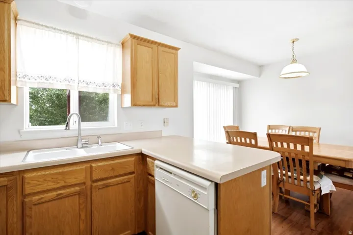 Wood cabinetry, white sink basin, dishwasher, light-toned countertops, and a dining area with a hanging light fixture