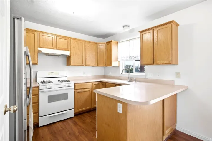 L-shaped kitchen with wood-finish flooring, light wood cabinetry, white appliances, a stainless steel refrigerator, and a double basin sink