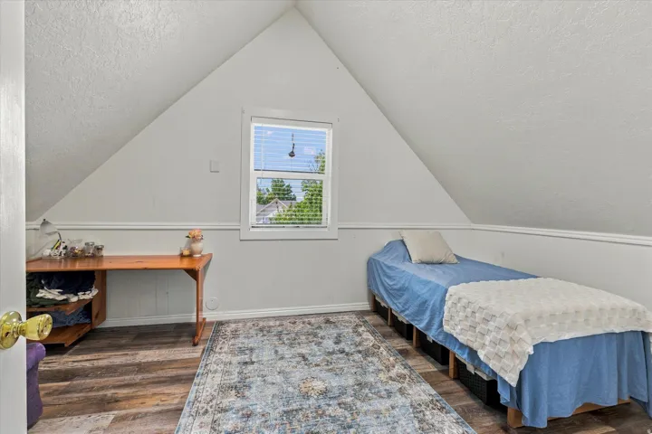 Sloped ceiling room featuring wood-finish flooring, white paneled lower walls, and a window with blinds