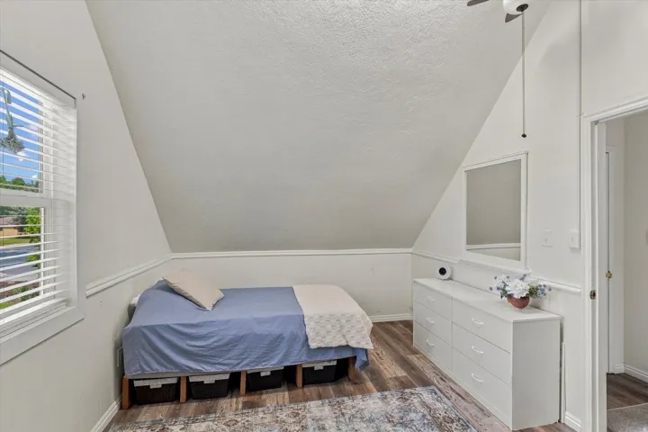 Angled ceiling room featuring wood-finish flooring, a window with blinds, and a ceiling fan