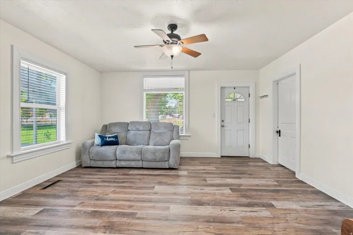 Spacious room with wood-finish flooring, multiple windows, a ceiling fan with integrated lighting, and a paneled entry door