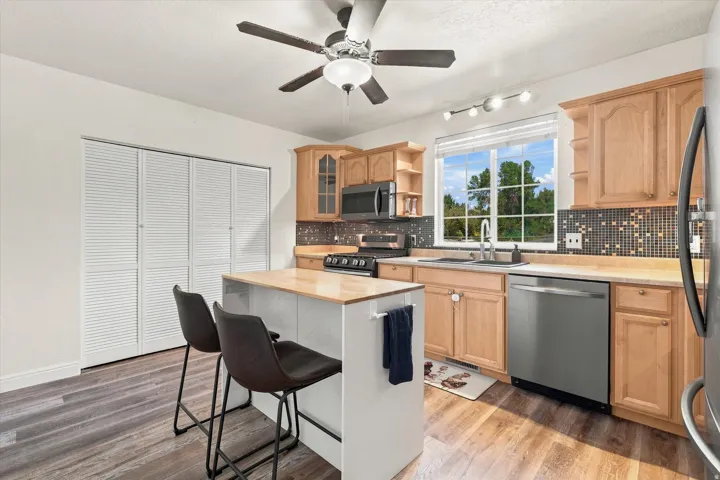 Kitchen featuring wood-finish flooring, light wood cabinetry, a mosaic tile backsplash, stainless steel appliances, and an integrated breakfast bar