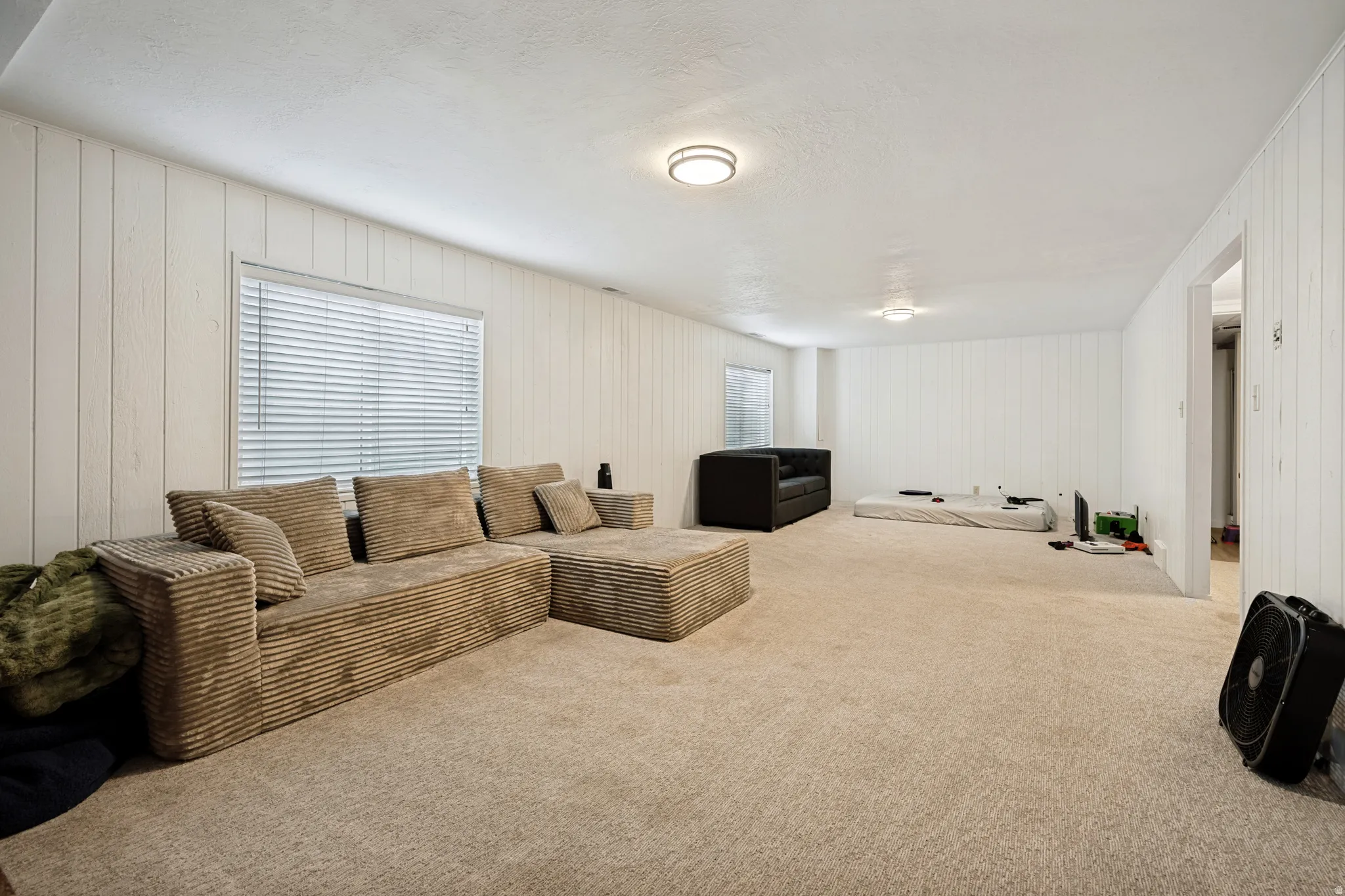 6711 - Basement Living Room. Spacious room featuring neutral carpeting, white paneled walls, a window with blinds, and flush-mount ceiling lighting