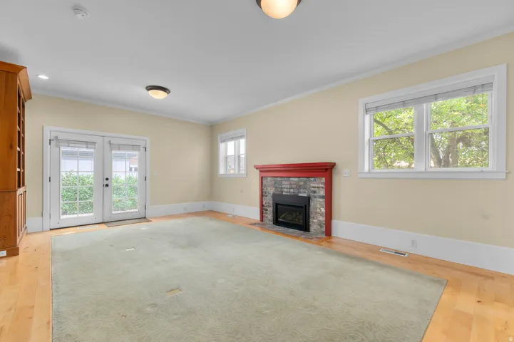 Wood-finish flooring, built-in wooden cabinetry, double glass-paneled doors, stone-faced fireplace with red mantel, and multiple windows