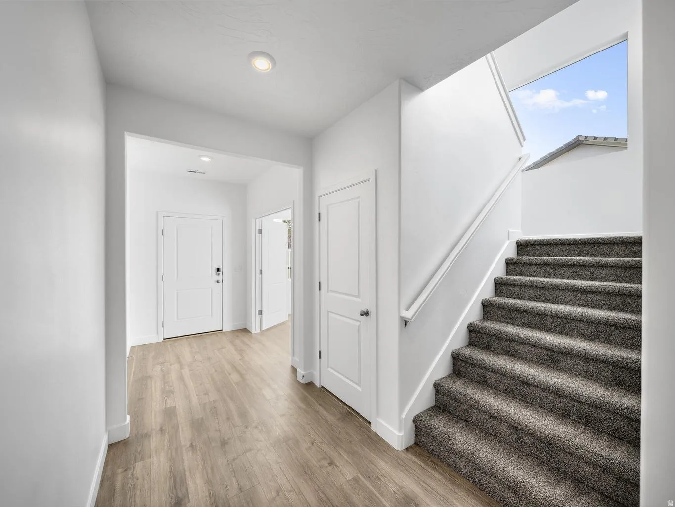 Light wood-finish flooring, white paneled doors, recessed ceiling lighting, and a carpeted staircase with a white railing