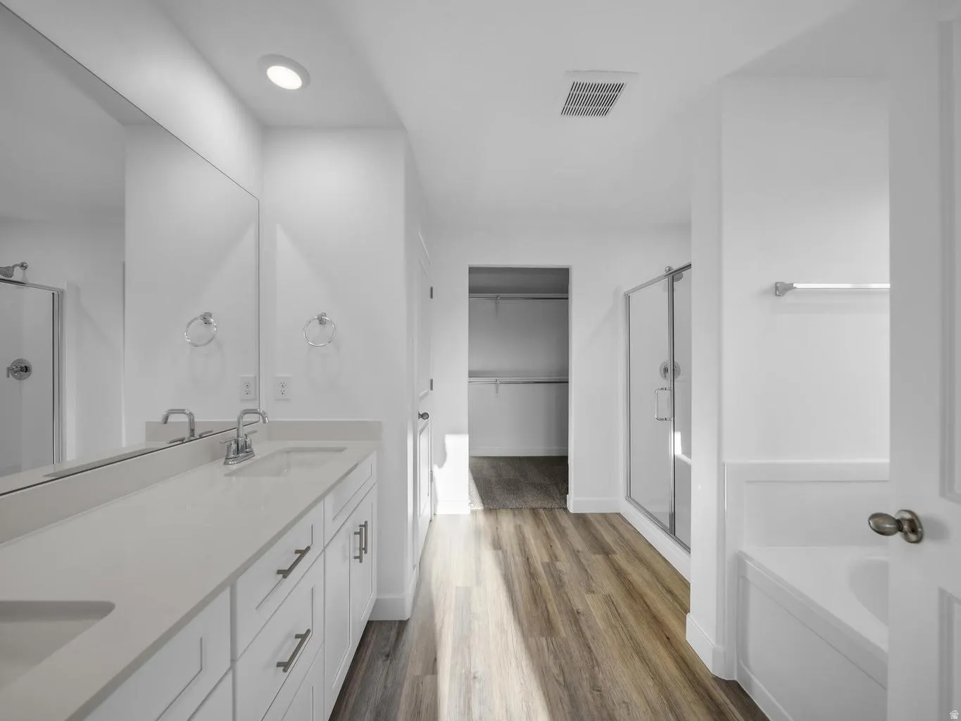 Dual vanity with integrated sinks and chrome fixtures, framed mirror, wood-finish flooring, glass-enclosed shower, and built-in bathtub