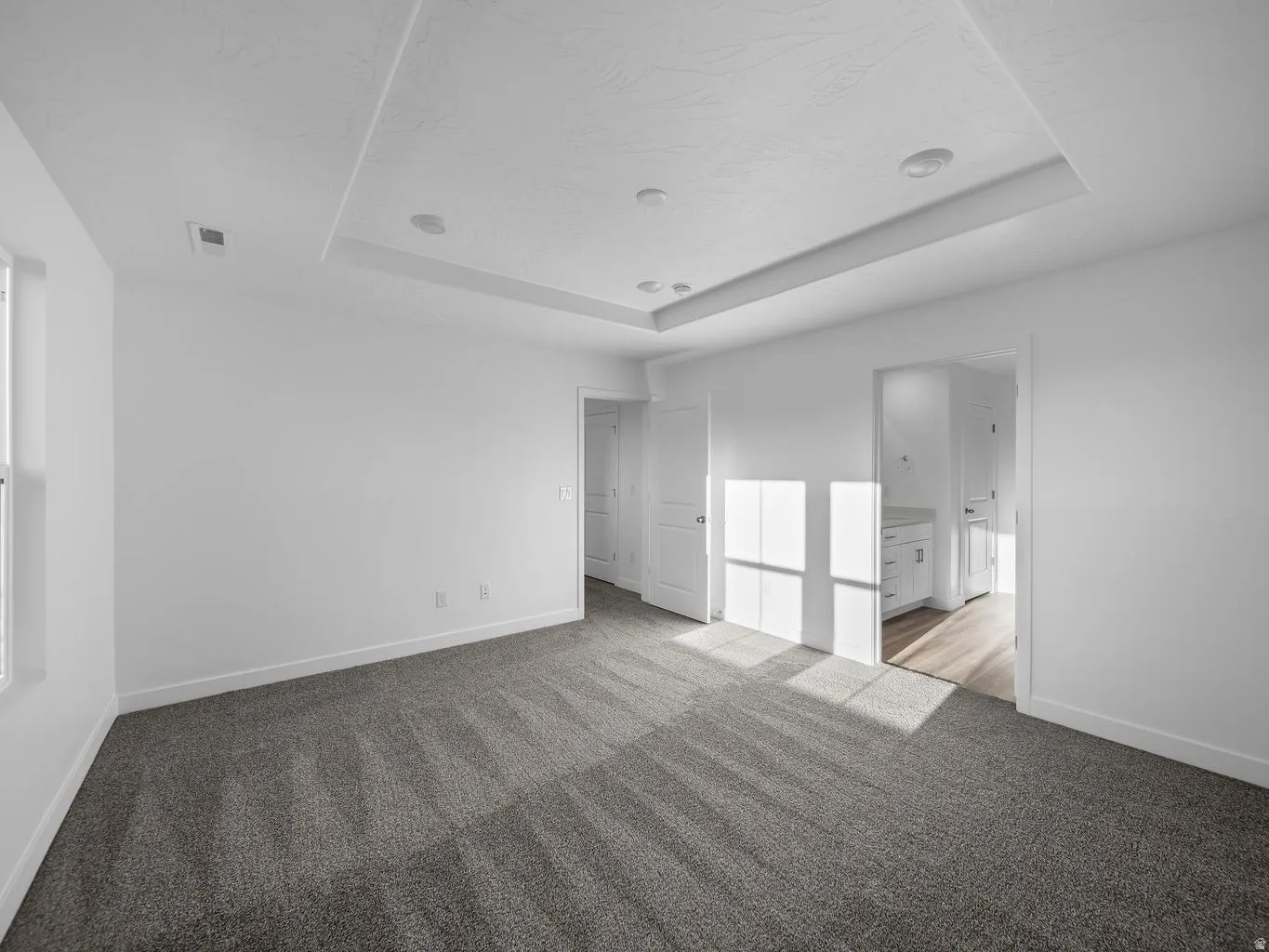 Recessed ceiling lighting, tray ceiling, neutral wall tones, carpeted flooring, and a window