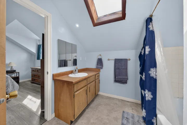 Sloped ceiling bathroom featuring a skylight, wood-finish vanity with an integrated sink, tri-fold mirrored medicine cabinet, and a tiled shower-tub combination