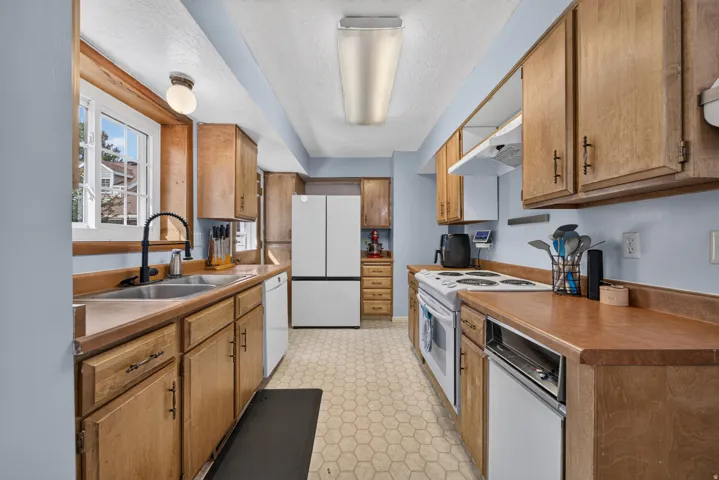 Galley kitchen featuring wood cabinetry, white appliances, double basin sink with a matte black faucet, and hexagonal floor tiling