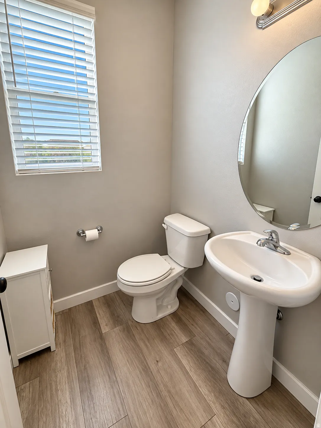 Pedestal sink with chrome fixtures, wood-finish flooring, and a window with horizontal blinds