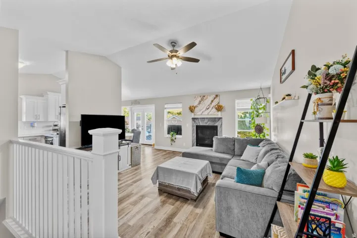 Vaulted ceiling with a ceiling fan, fireplace with a stone surround, wood-finish flooring, white railing, and white kitchen cabinetry
