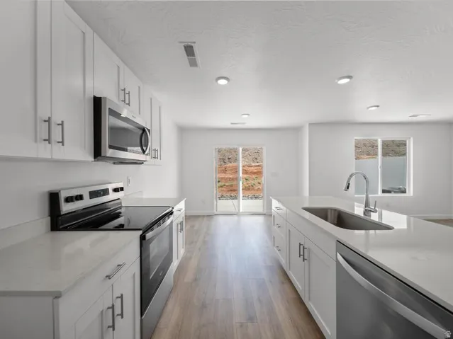 Kitchen featuring white cabinetry, stainless steel appliances, light-toned countertops, a kitchen island with an integrated sink, and wood-finish flooring