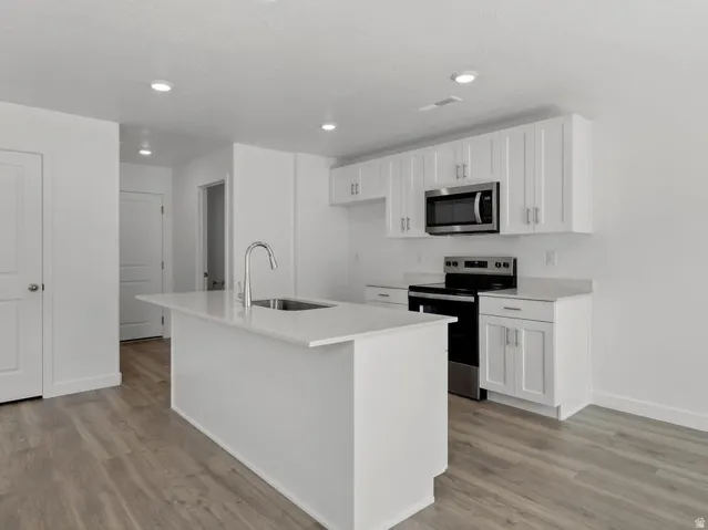 Kitchen featuring an island with an undermount sink, stainless steel appliances, white cabinetry, and wood-finish flooring