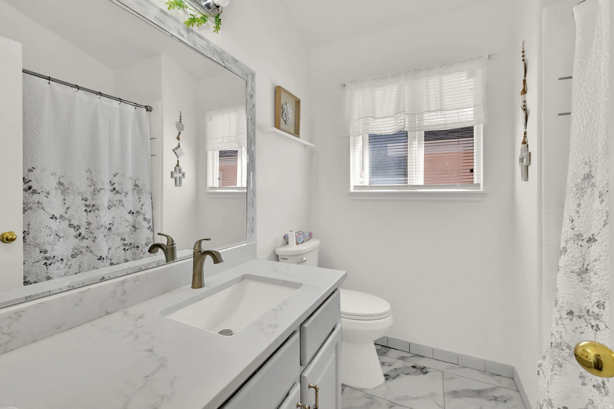Vanity with integrated sink and bronze-finish faucet, large framed mirror, toilet, and window with blinds