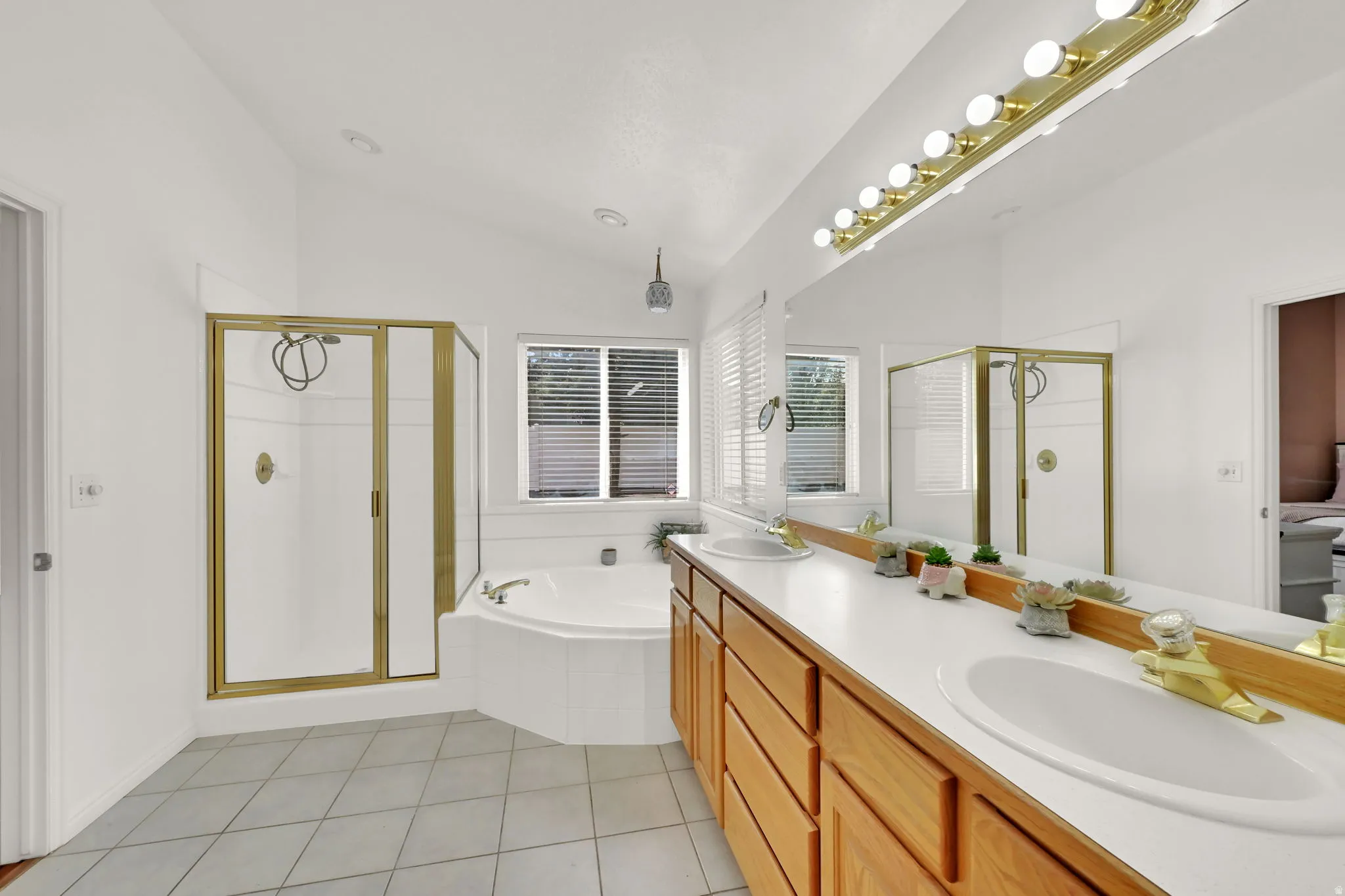 Dual vanity with wood cabinetry, large soaking tub, separate shower with brass-finish enclosure, tile flooring, and vaulted ceiling