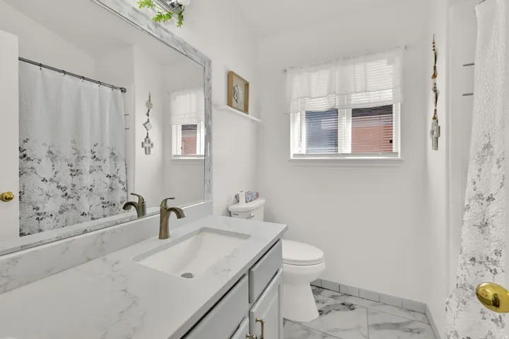 Vanity with integrated sink and bronze-finish faucet, large framed mirror, toilet, and window with blinds