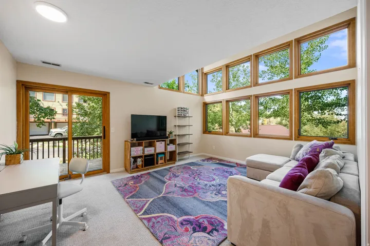Lower level second living room with deck to front of the house. Vaulted ceiling with a flush-mount light fixture, a wall of windows with wood-finish frames, a glass sliding door with wood-finish frame, and carpet flooring