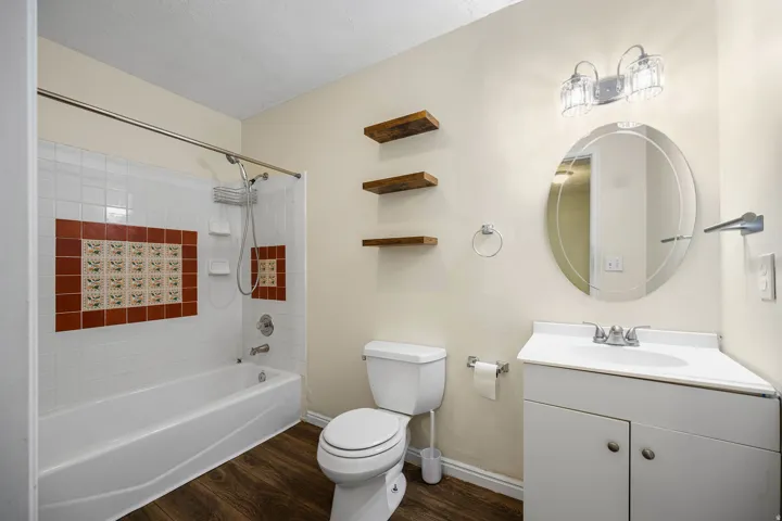 ADU full bathroom connected to primary bedroom. Wood-finish flooring, white vanity with integrated sink, circular wall mirror, and a tub with decorative tiled surround