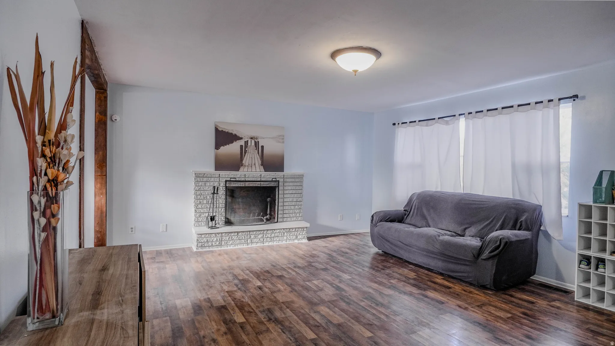 Wood-finish flooring, white brick fireplace, multiple windows, and a flush mount ceiling light