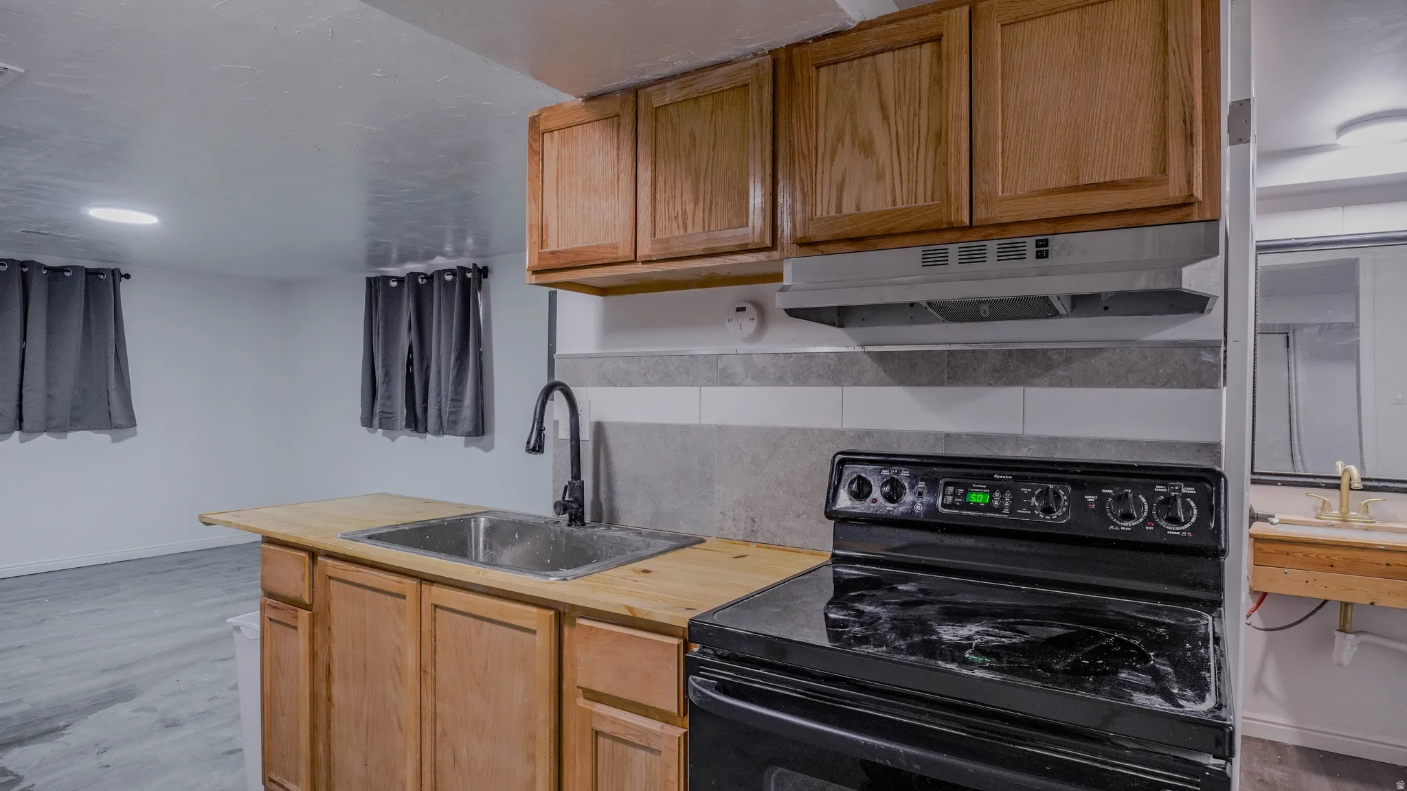 Kitchenette featuring wood-finish cabinetry, a stainless steel sink with a matte black faucet, a black electric range, and a tiled backsplash