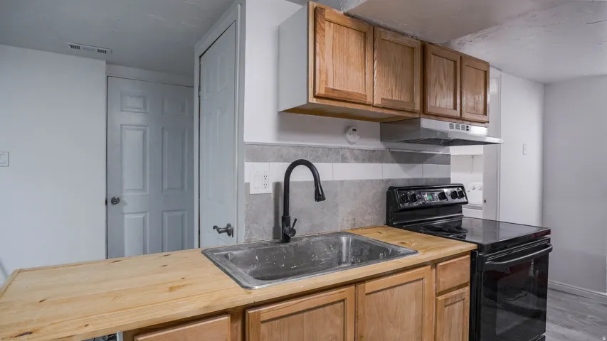 Kitchenette featuring wood-finish cabinetry, stainless steel sink with matte black faucet, electric range, and range hood