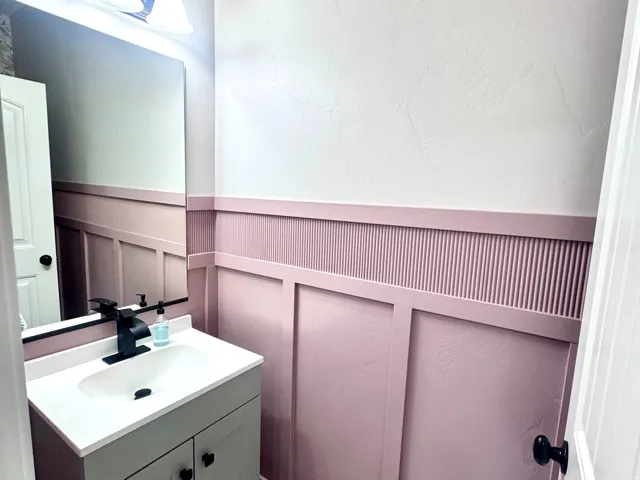 Vanity with integrated sink, black matte faucet, wall-mounted light fixture, decorative wall paneling, and tile flooring
