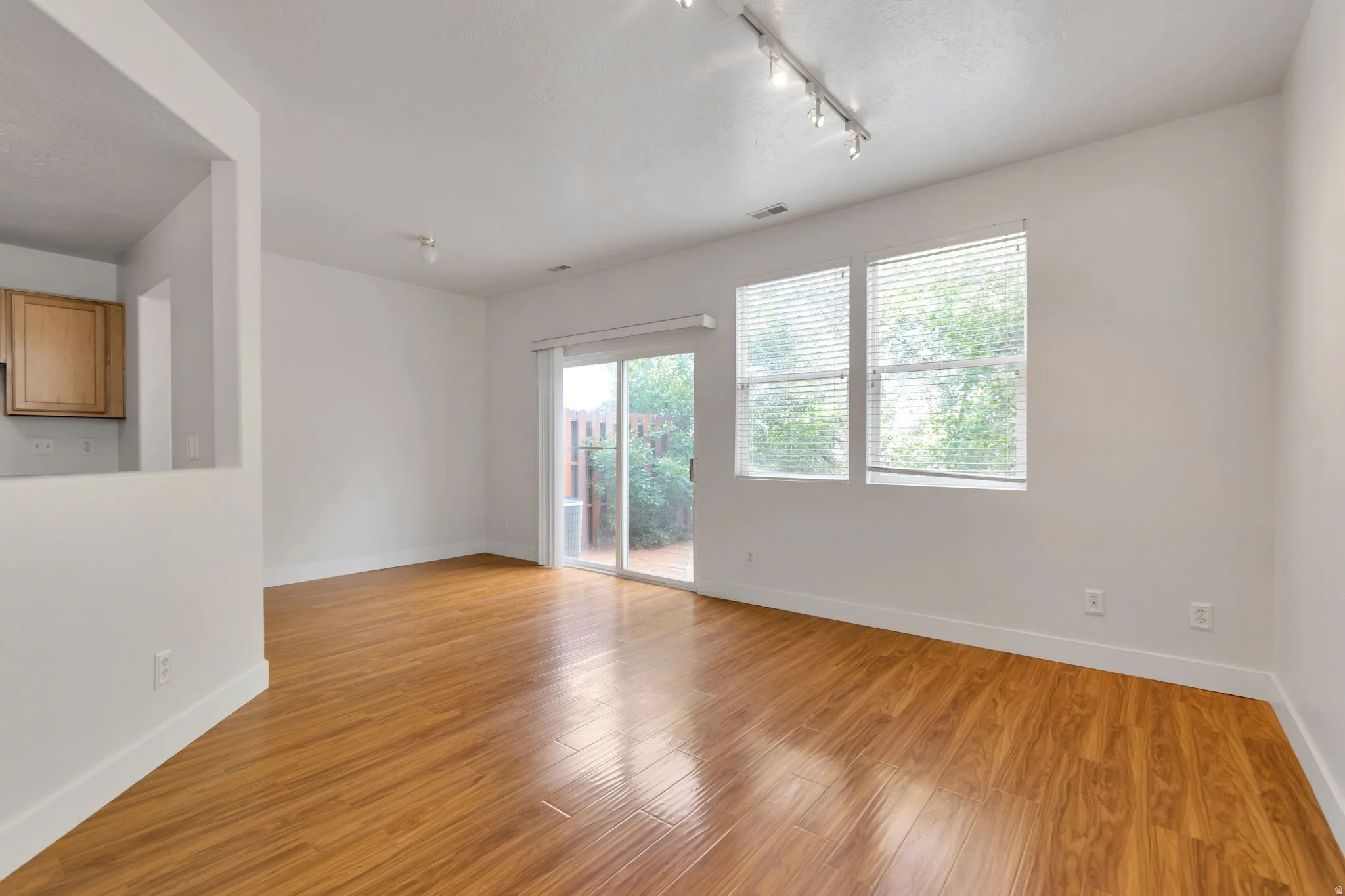 Wood-finish flooring throughout, multiple windows, track lighting fixture, sliding glass door, and a partial wall with built-in cabinetry visible