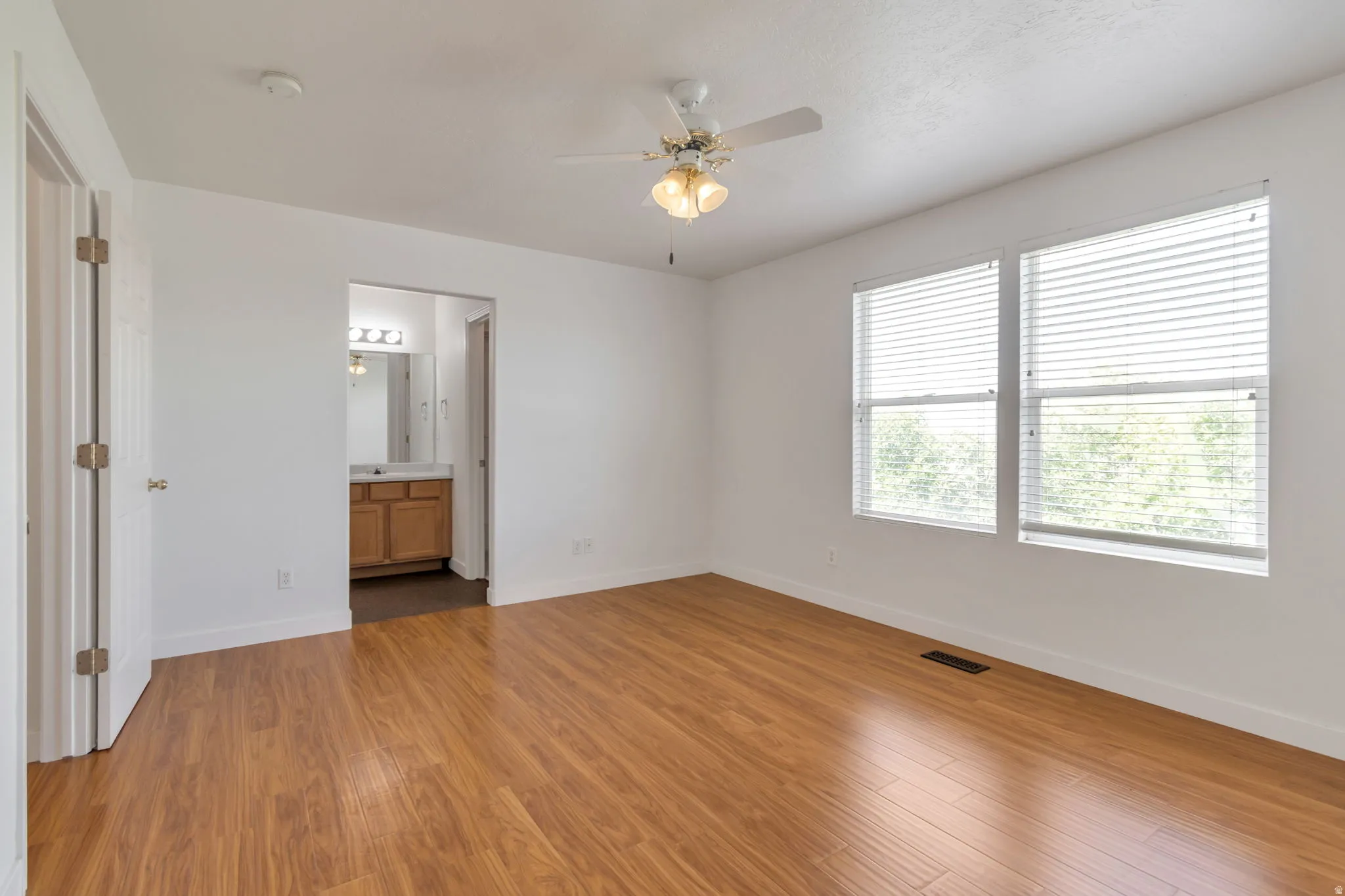 Spacious room featuring wood-finish flooring, two large windows with blinds, and a ceiling fan with integrated lighting