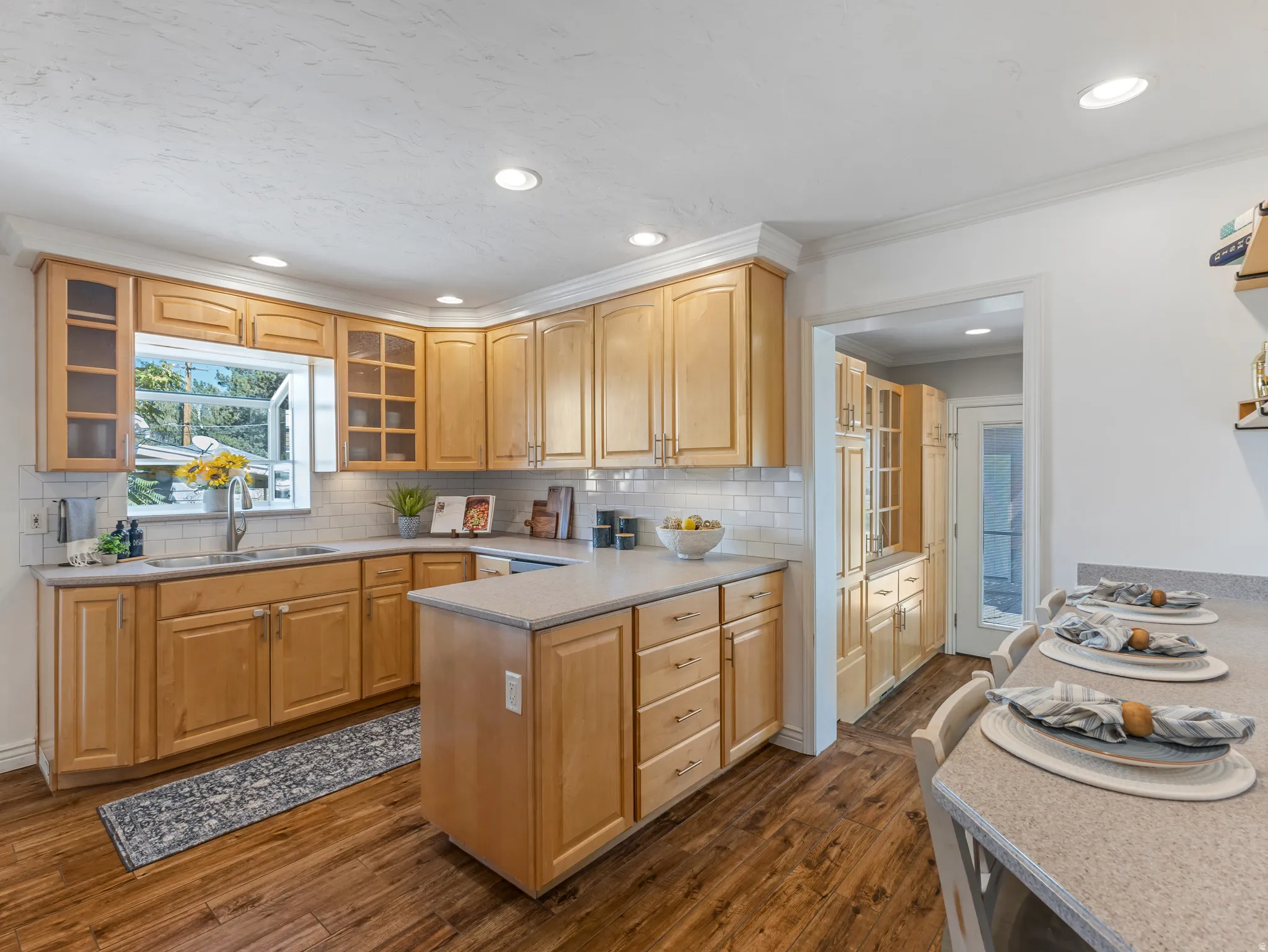 Wood-finish flooring, light wood cabinetry with glass-front accents, white subway tile backsplash, recessed lighting, and a kitchen island with countertop seating