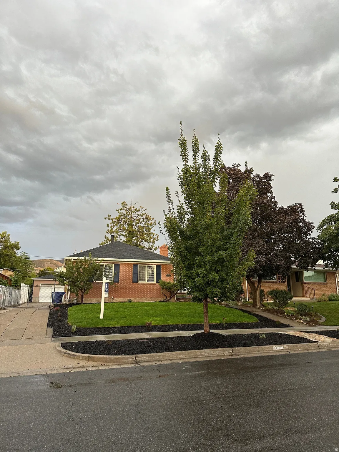 Red brick exterior with dark shutters, asphalt shingle roof, paved driveway, and established front yard landscaping
