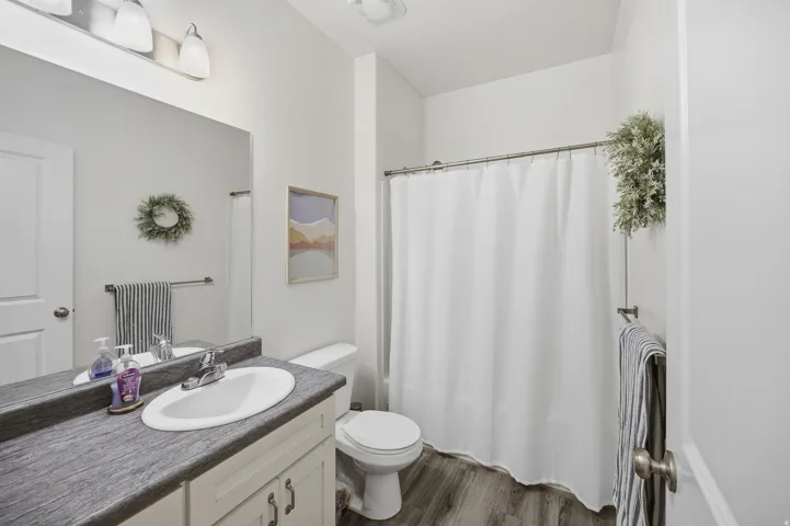 Vanity with an integrated sink, a large wall-mounted mirror, wood-finish flooring, a white toilet, and a white shower curtain rod