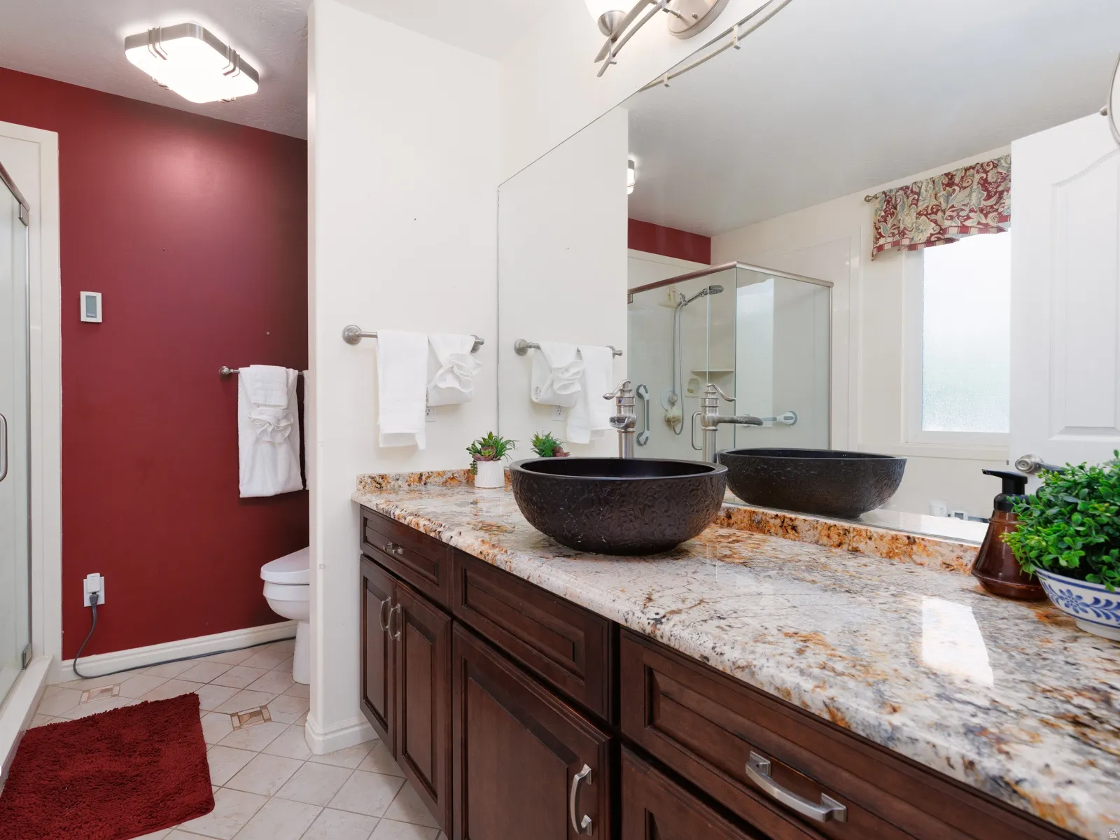 Dual vessel sinks set in an expansive vanity with a light-toned countertop, a glass-enclosed shower, and tile flooring